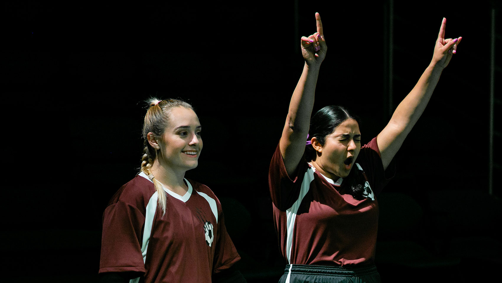 Two women's soccer teammates stand near each other, one cheering as she raises both arms upwards in victory signs, the other smiling as she looks on whatever is happening before them.