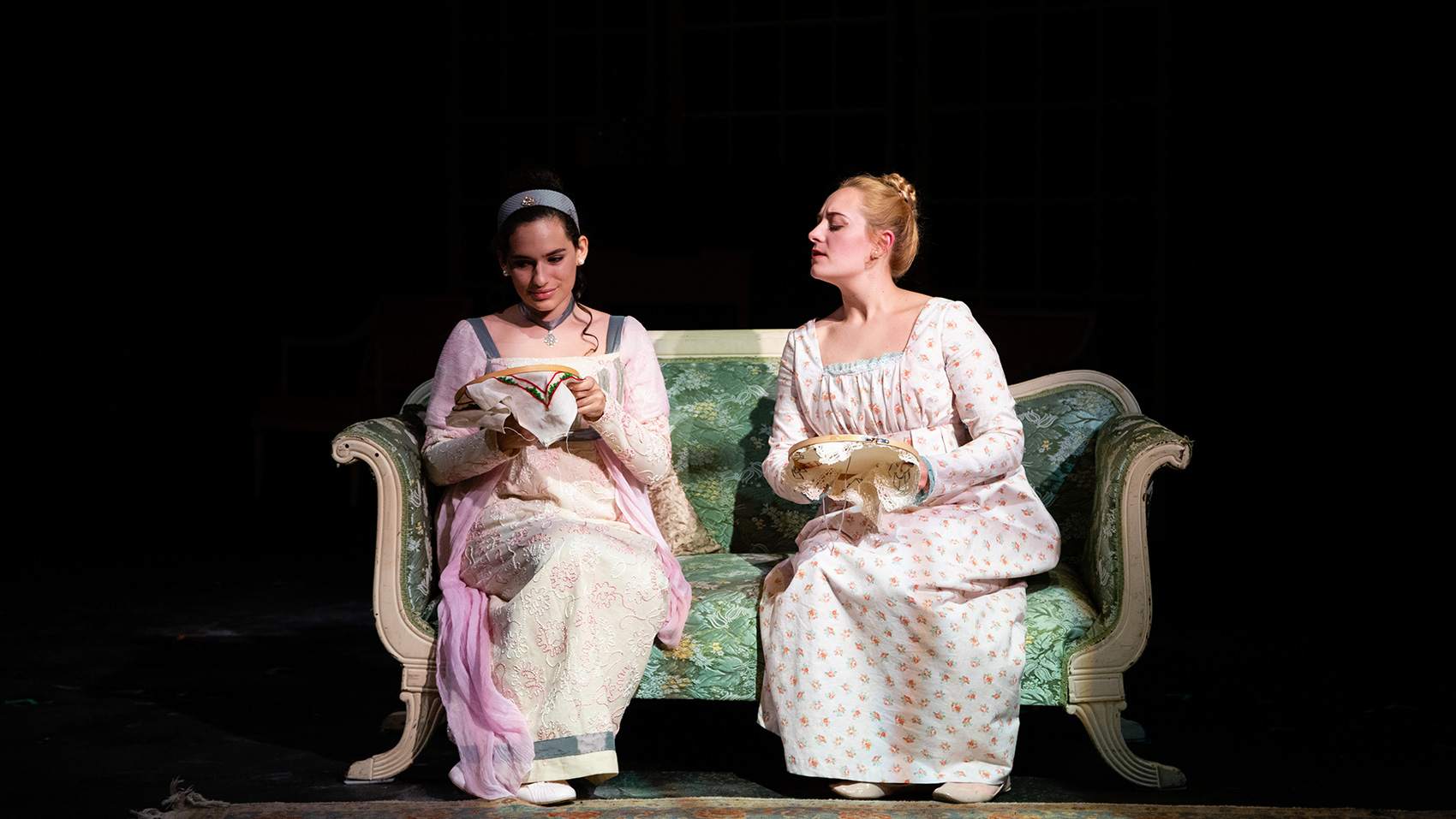 Two women sitting and cross stitching on a couch, one looking over curiously at what the other is doing.