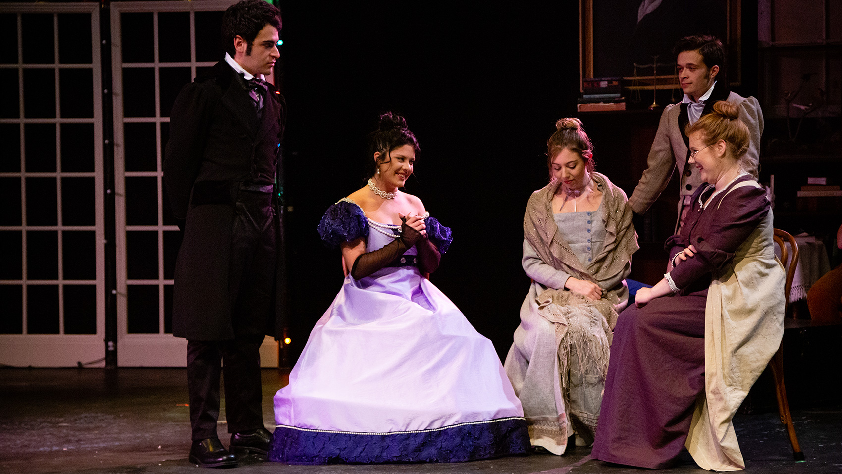 Three women sitting in a circle facing each other, talking excitedly. Two men stand with them, looking on.