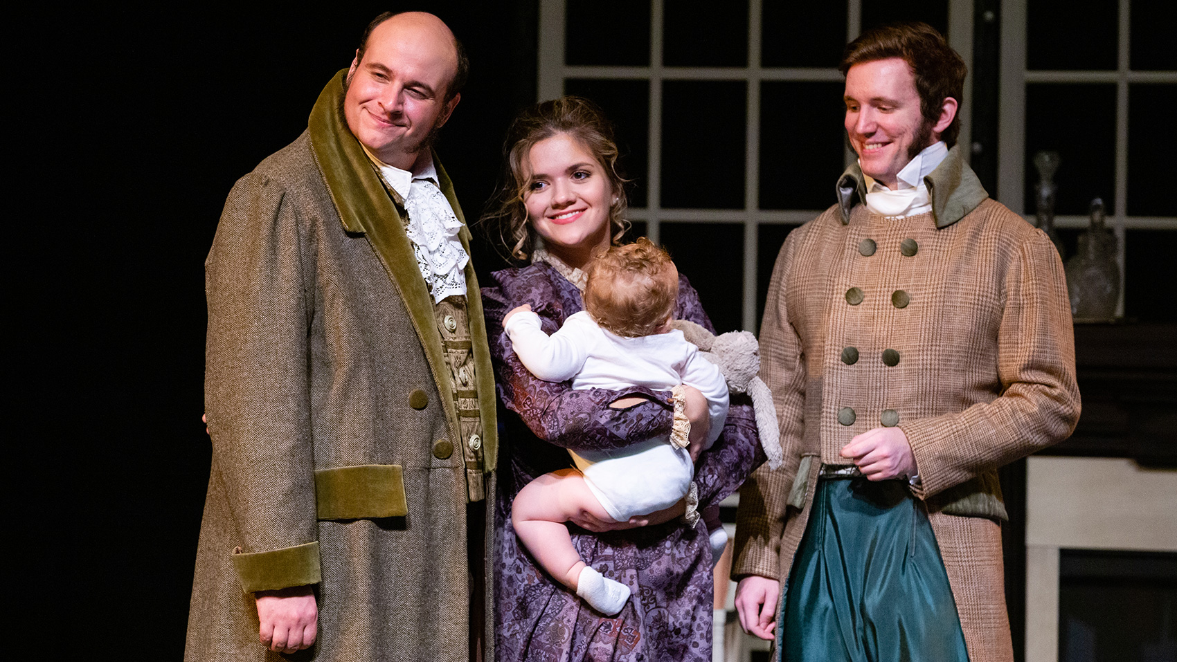 A smiling woman holds a baby, two men stand with her, one of each side.