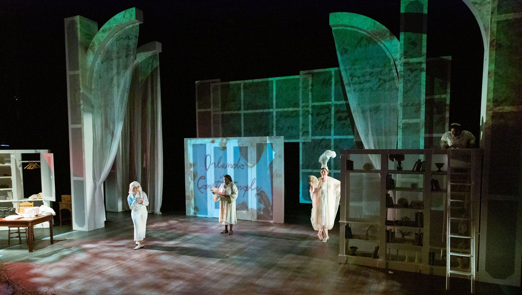 Three people stand far apart from each other in a room, the stage is lit up with white and green lighting. All three people wear white clothing, 