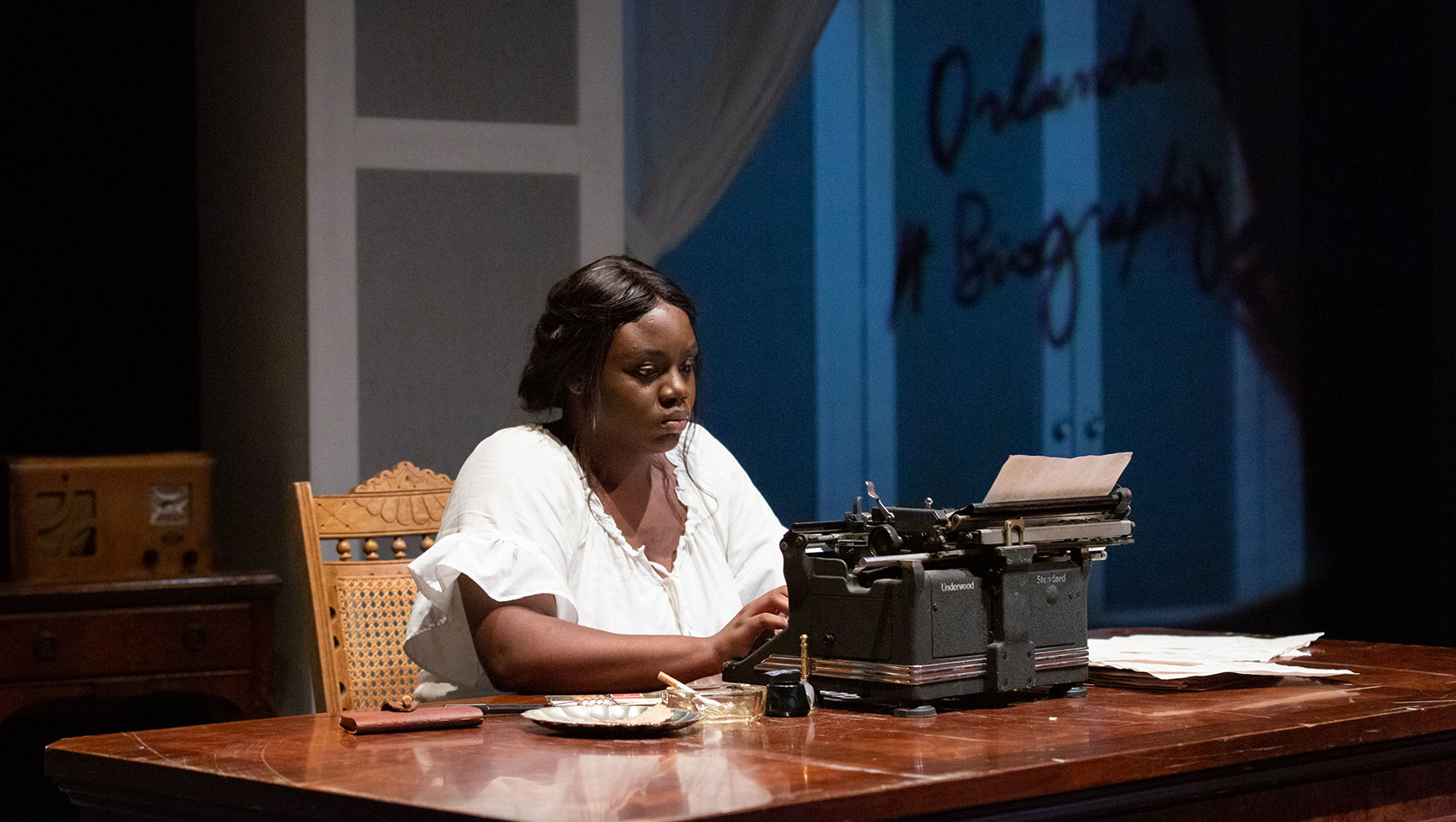 A Black woman sits alone typing something using a typewriter, her face serious as if she is thinking carefully. 