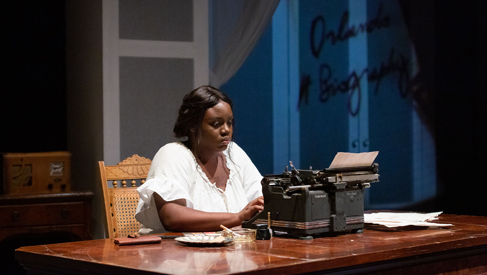 A Black woman sits alone typing something using a typewriter, her face serious as if she is thinking carefully.