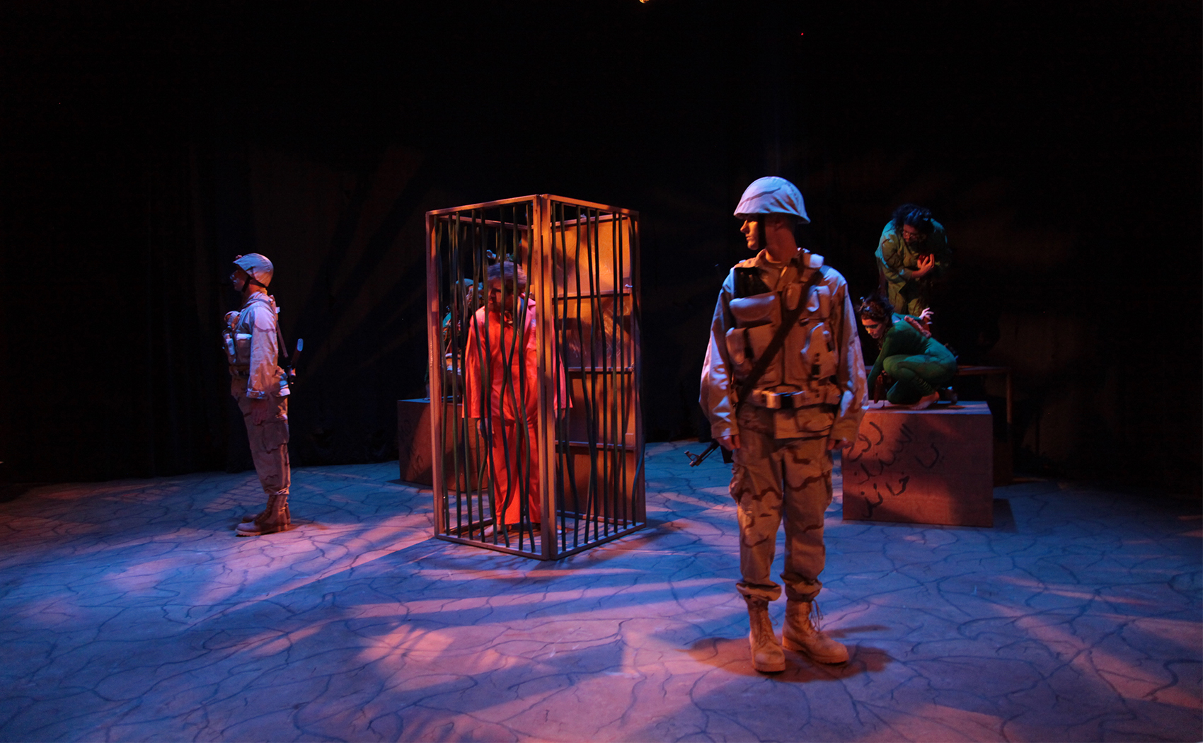 A man is hunched over, decked in an orange jumpsuit, looking scornfully. Several soldier guards stand watch around his cage, armored and geared. Some boxes with a foreign language scrawled across it are in the back, two people dressed in green crouch on them.  