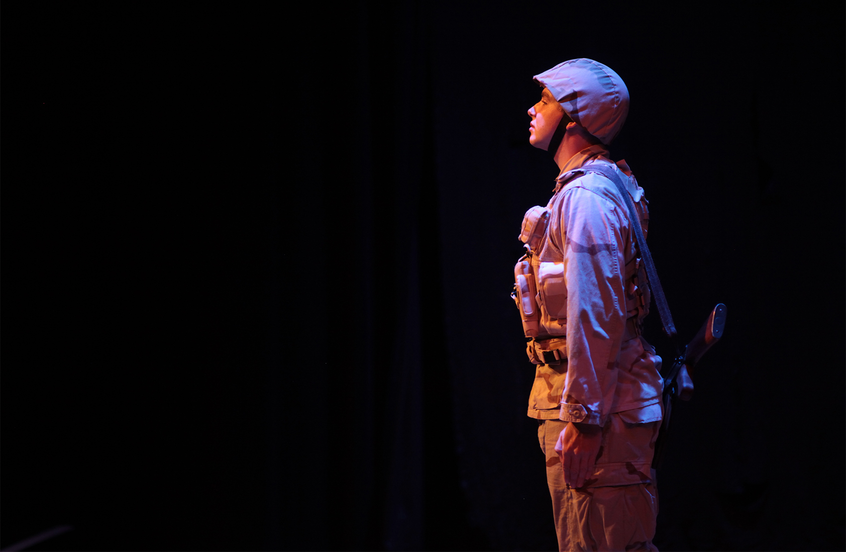 A young military solider stands in his helmet and accompanying gear, rifle against his back as he looks out and in silence or reverie. The background is dark against his light figure.  
