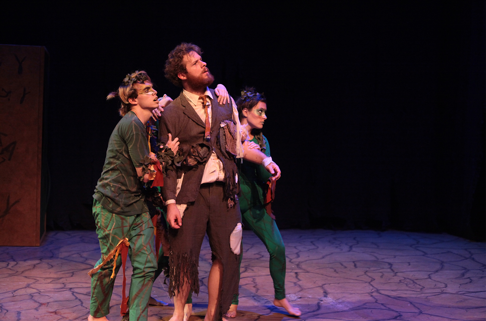 Two figures (one young man and one young woman) dressed in colorful green clothing, feathers and miscellaneous items scattered on their form and in their hair stand side by side against a man with a rugged beard in a tattered suit. The three of them are tensed, anticipating whatever might be coming from off-screen.  