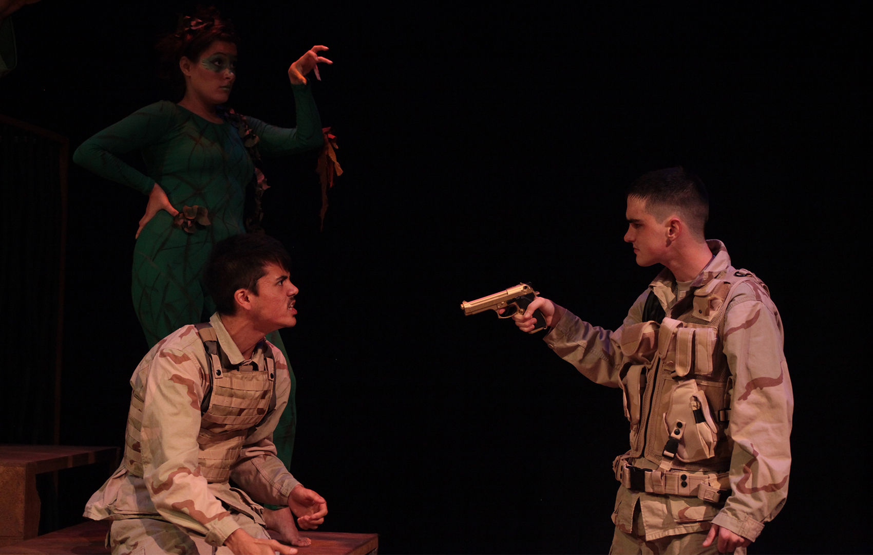 This scene showcases the middle of an intense argument/confrontation. A young woman dressed in green clothing stands in the background, shadowed by darkness, her hand posed in a clawing manner. She stands in the teapot position. In the foreground, two young men are faced to each other, one sitting down yelling at the other one, who stands with a gun pointed at the former. Both are wearing military gear.     