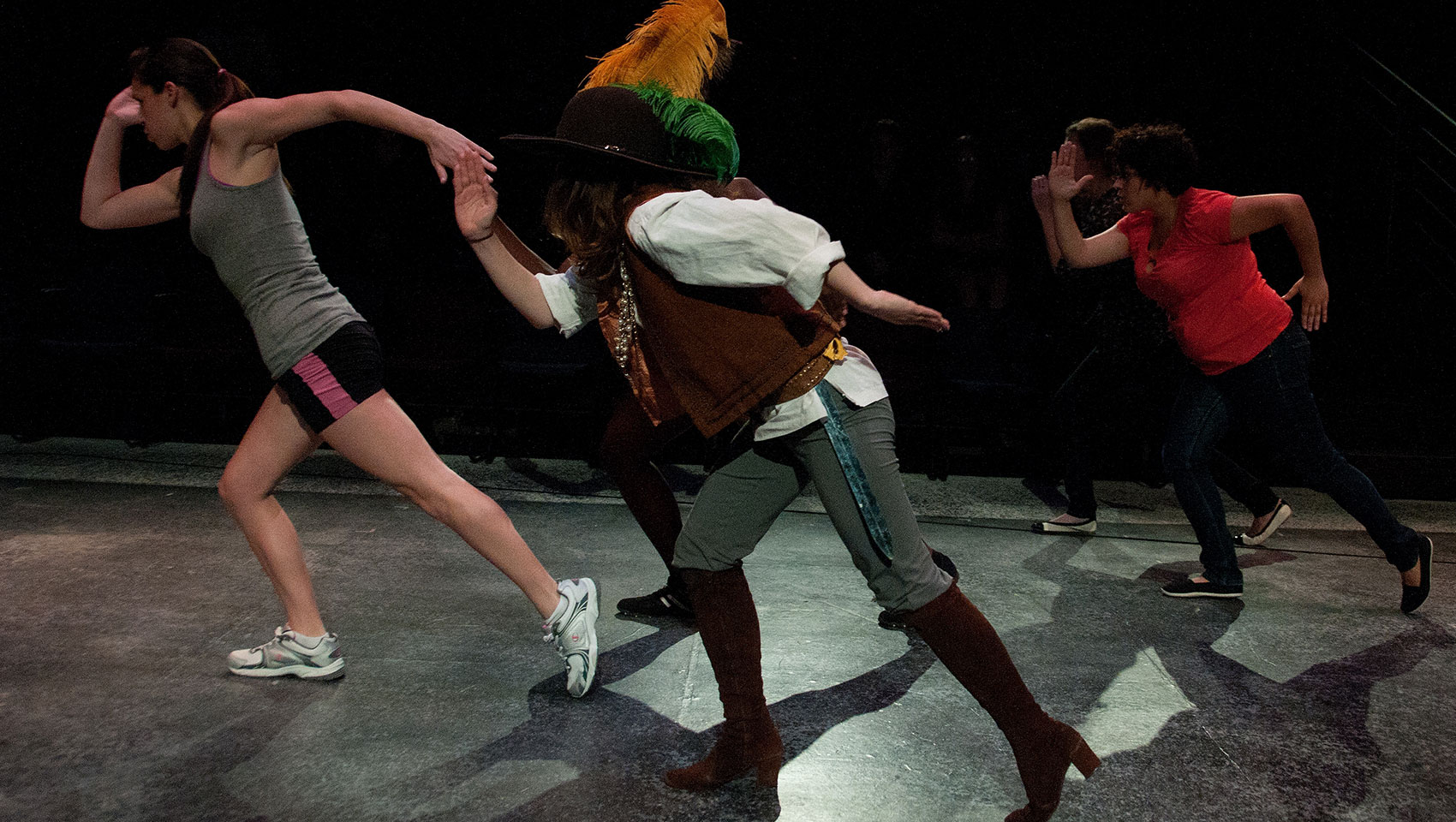 Four people hold the same pose as if in mid-run. They raise their right arm, elbow bent 90 degrees, and their right leg lunges forward. All except one appear to be wearing exercise clothes. The one outlier wears a hat with two large feathers attached, and a brown vest over a white button-up, as well as knee-high heeled boots. 