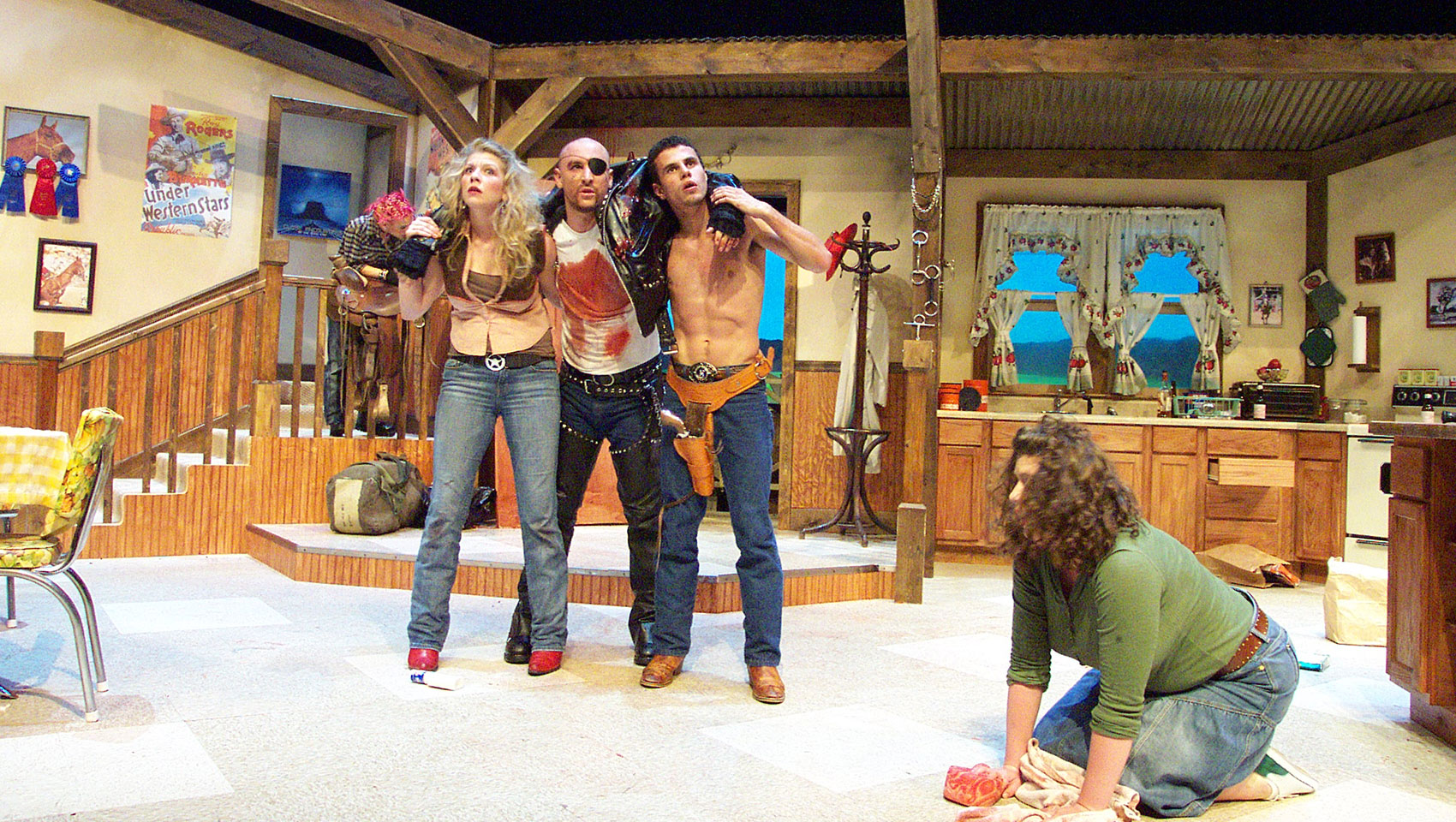 An injured man with a bloodied t-shirt and an eye patch walks with the help of another man and a woman, his arms draped over them. Yet another woman kneels on the floor in front of them, her face hidden by her hair. She holds onto bloodied cloth.