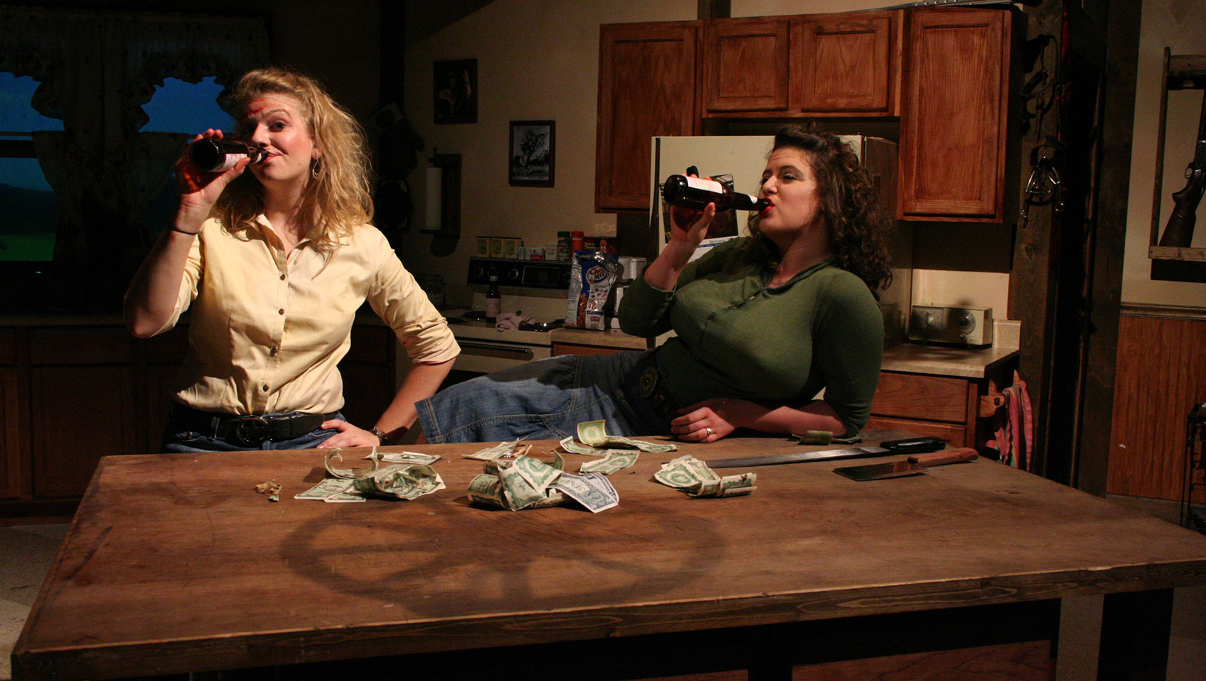 Two women relax in the kitchen drinking beer, one standing and the other sitting on a stool and leaning against the table. 