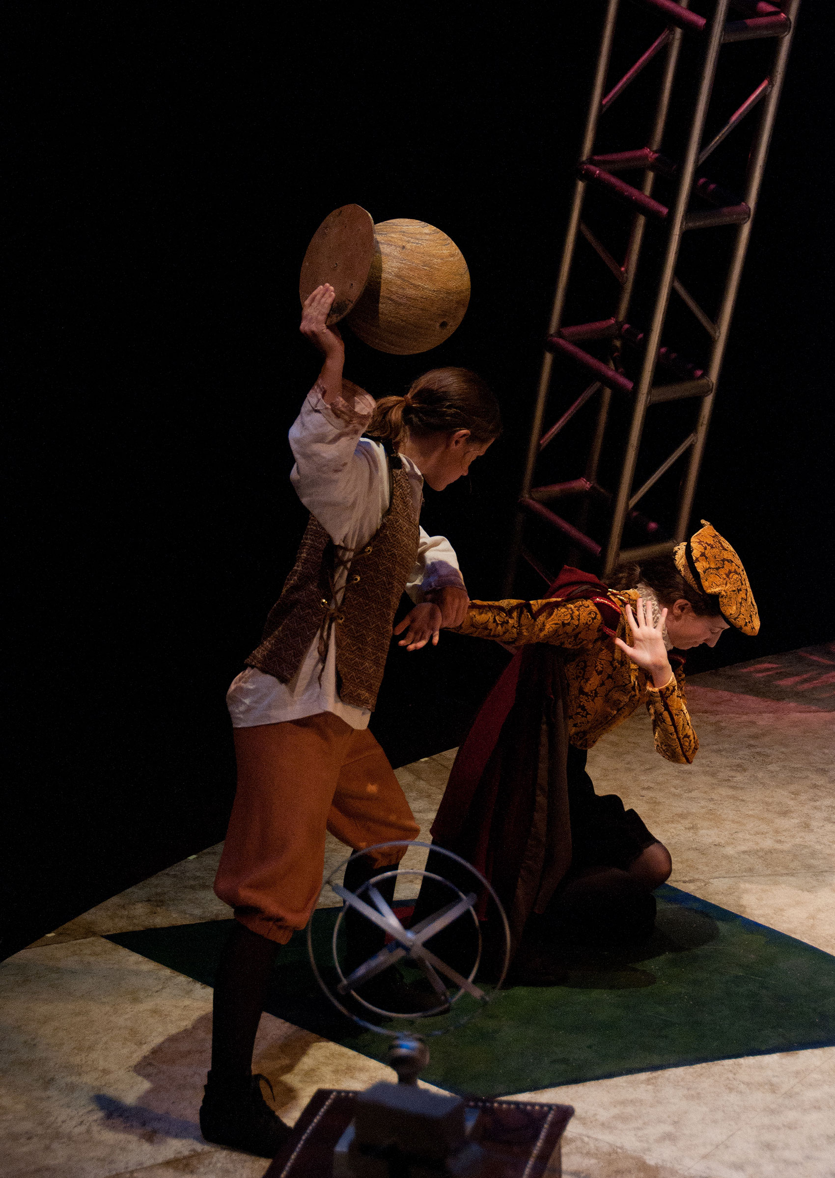 A man kneels on the floor and tries to shield his face with one hand from another person who is threatening to hit them with a large round object. 