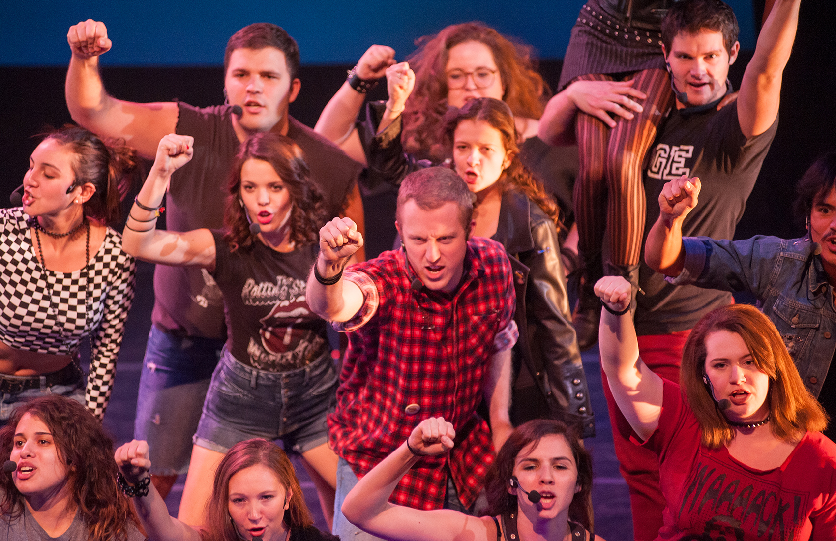 A crowd of young men and women decked in red and black alternative punk styled clothing are crowded around one another, one fist punched in the air as they look at the audience with a strong and defiant focus.