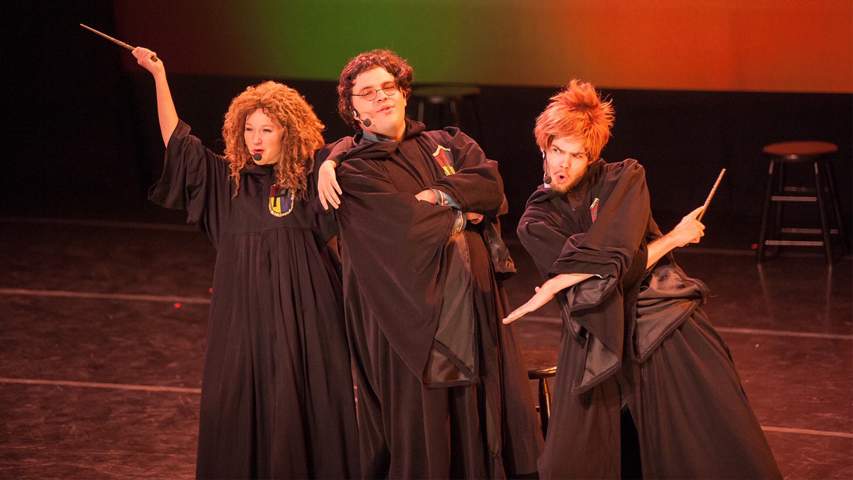 Three actors dressed as characters from the Harry Potter series stand together and sing. On the far left, the actress holds up a wand and has her arm against the shoulder of the central actor, who leans against her. The right actor has his arms crossed in an ‘X’, wand in one hand.  