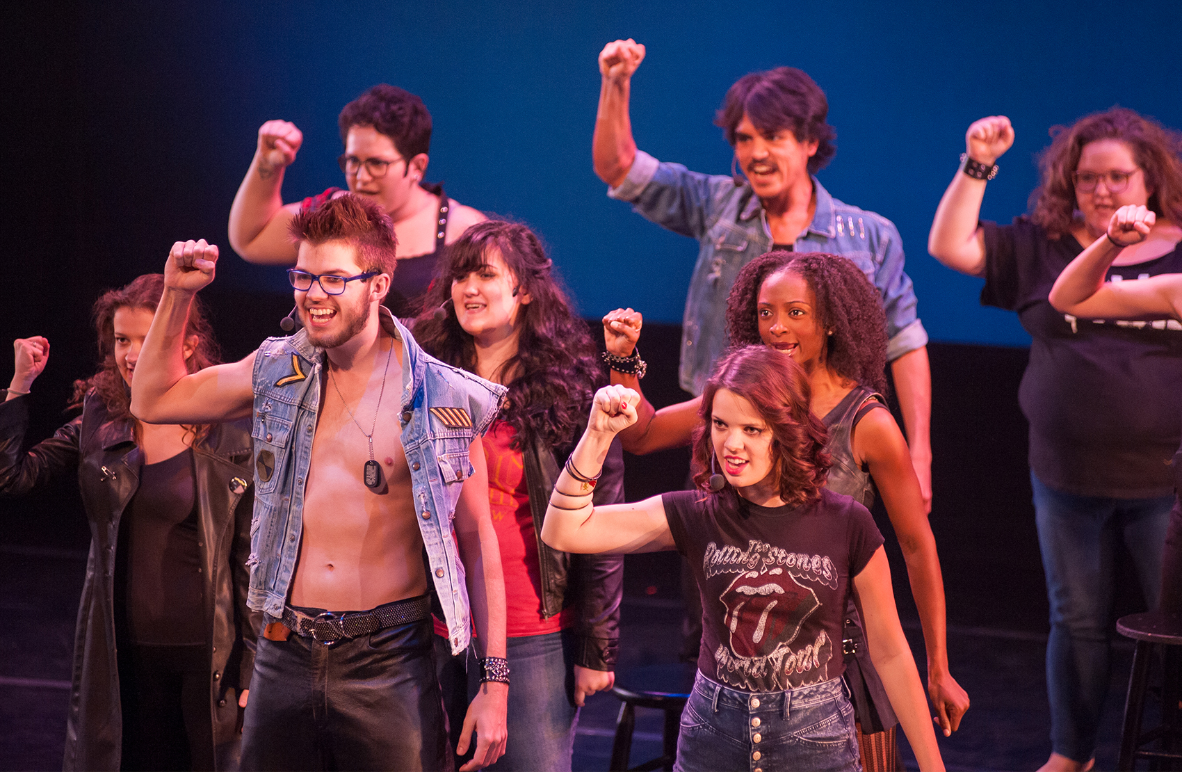    Another crowd of young men and women, dressed in alternative punk dark and denim clothing, sing toward the audience and have their right fists raised, arms at a 90-degree angle.  