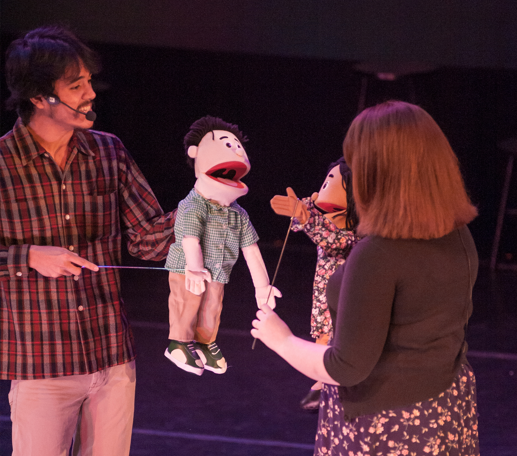 The camera is angled from above, and its placed on a young man and the back of a woman. Each of them holds a puppet dressed in a similar fashion to their outfits: the woman decked in a floral dress and a black cardigan, the man in a plaid long sleeve shirt and khaki pants. 