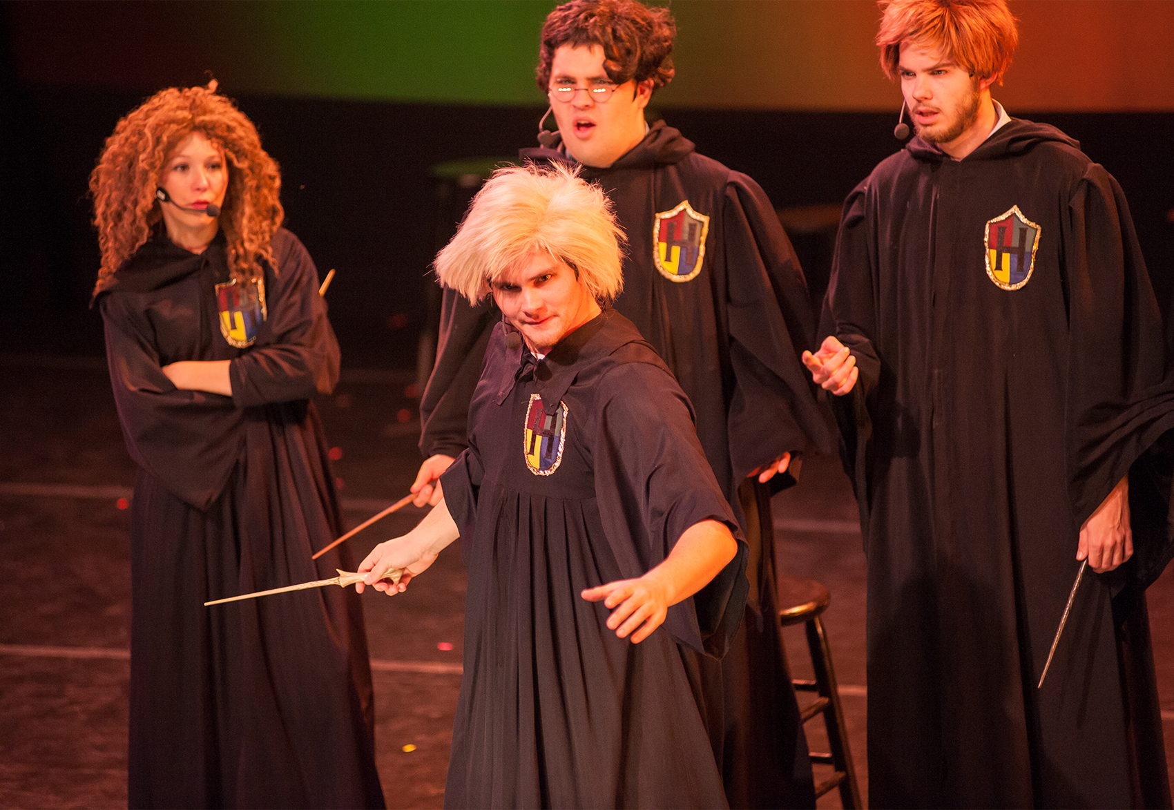 A group of actors dressed as characters from the Harry Potter series stand huddled around one actor who stands in the foreground. He has his wand in his right hand and looks mischievously at the audience. The actress behind him to the right dons a curly headed wig and has her arms crossed, looking at the central actor. Next to her in the center background is an actor with glasses and curly hair who looks confused, along with the actor on his left, a wand in his left hand.