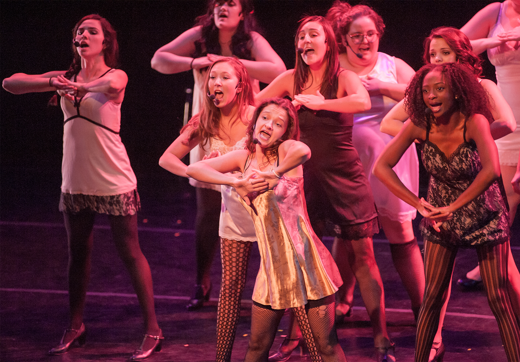 A group of young women clad in silken lace dresses and dark stockings sing toward the crowd, elbows pointed outward and hands either clasped together or placed over one another stretched out.