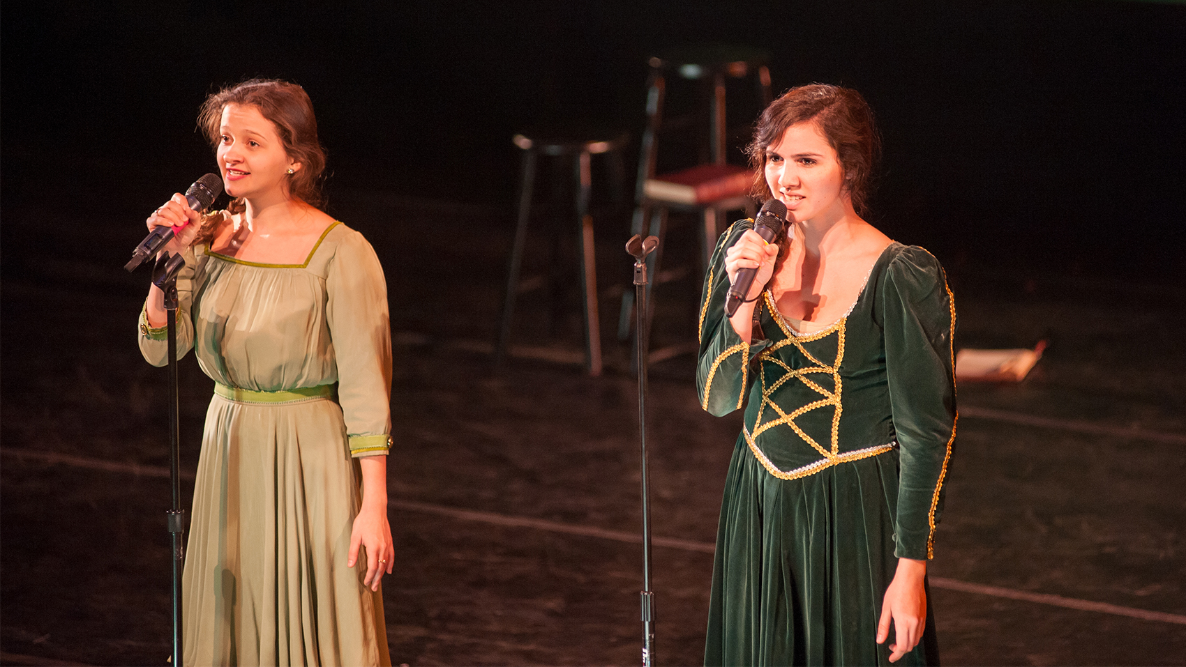 Two women in a beige vintage dress and a darker green and golden embroidered dress sing toward an audience. The girl on the left is holding the microphone in the stand, the other is holding the microphone off the stand.