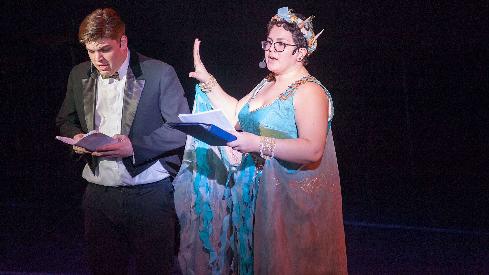 A young man in a tuxedo looked hurried, reading off of a script. A young woman in a seashell crown and blue flowy dress and cuffed glittery wings holds a blue binder and script in her left hand, her right pressed outward in a stopping motion at the young man standing beside her.