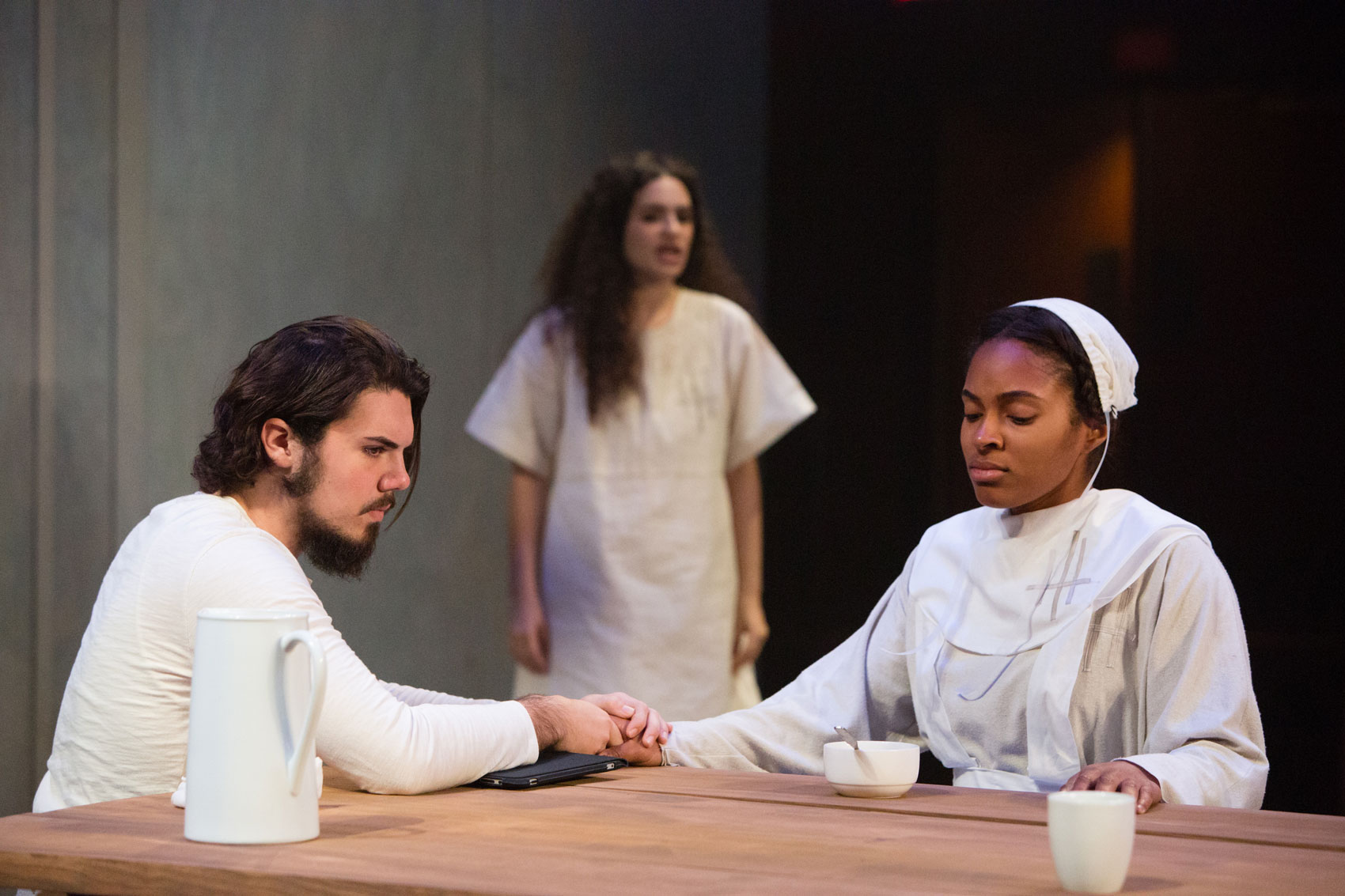 A man holds a woman's hand at a table while another woman looks on in the background, seemingly unnoticed.