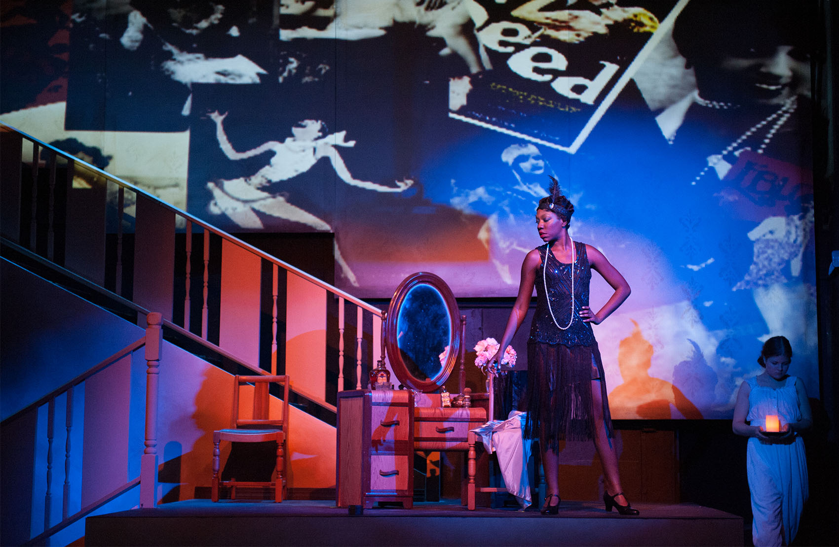 A young black woman in a sequin black flapper dress and matching feather headpiece glances downward, hand on her hip, right foot turned outward. Right next to her is a small wooden vanity, a small chair sits off to the side and a staircase is off to her left. The background/sky above her is dotted with photos of the past: flapper girls, vintage advertisements, jewelry. To her far right stands a young girl in pajamas, she looks forlorn and holds a glowing amber candle.