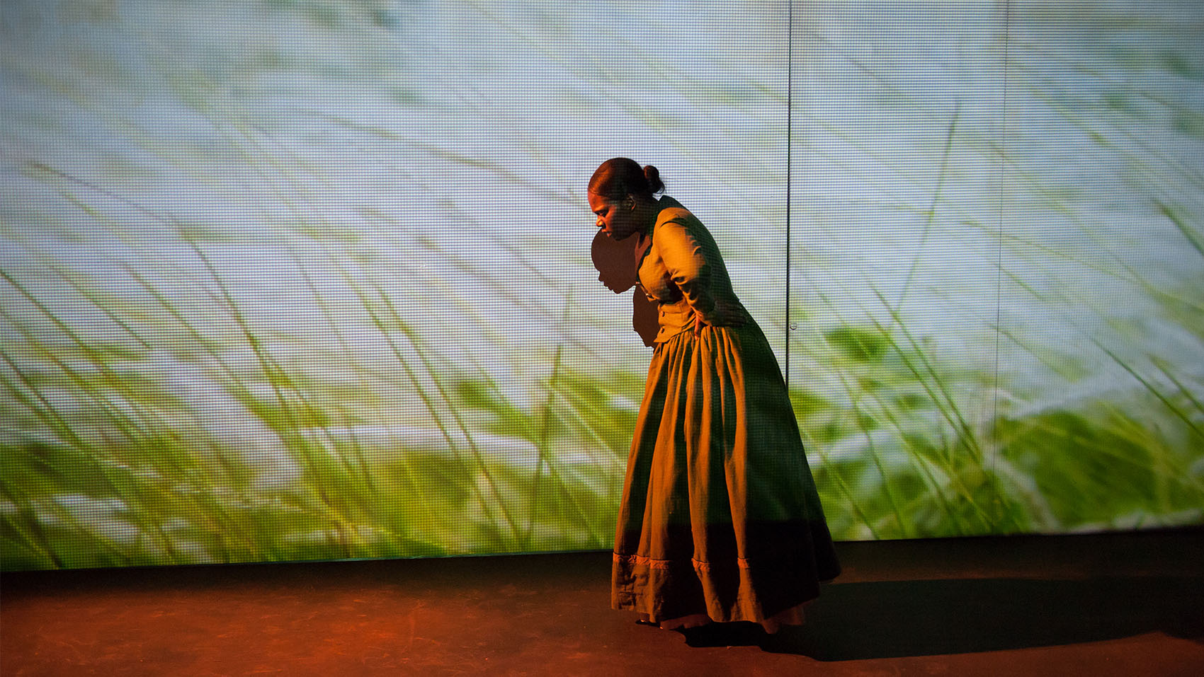 A young black woman in a vintage dress looks out of breath, her hands on her hips a screen projected with field crops and grasses stands behind her.