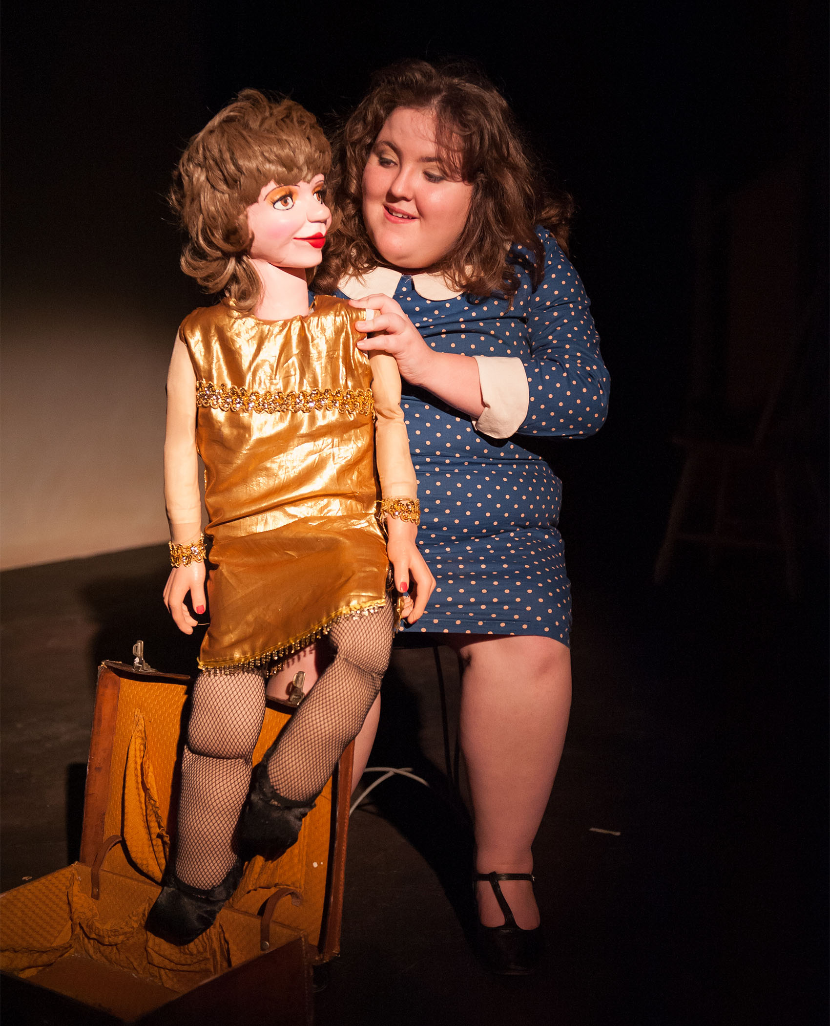 A ventriloquist sits atop a stool, in a blue cuffed and collared polka dot dress. Her puppet dummy sits on her knee, the puppet wears a gold shiny dress, a similarly colored suitcase sits beside her foot.
