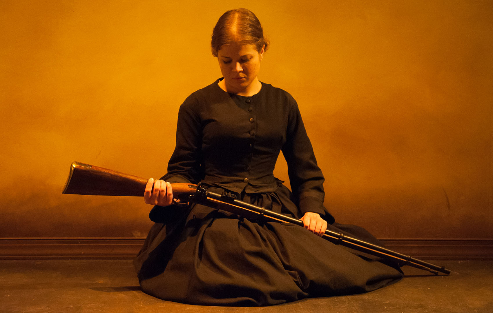 A young white woman is cast in deep yellow lighting, wearing an all-black vintage dress. She looks deep in thought and holds a long barrel rifle. 