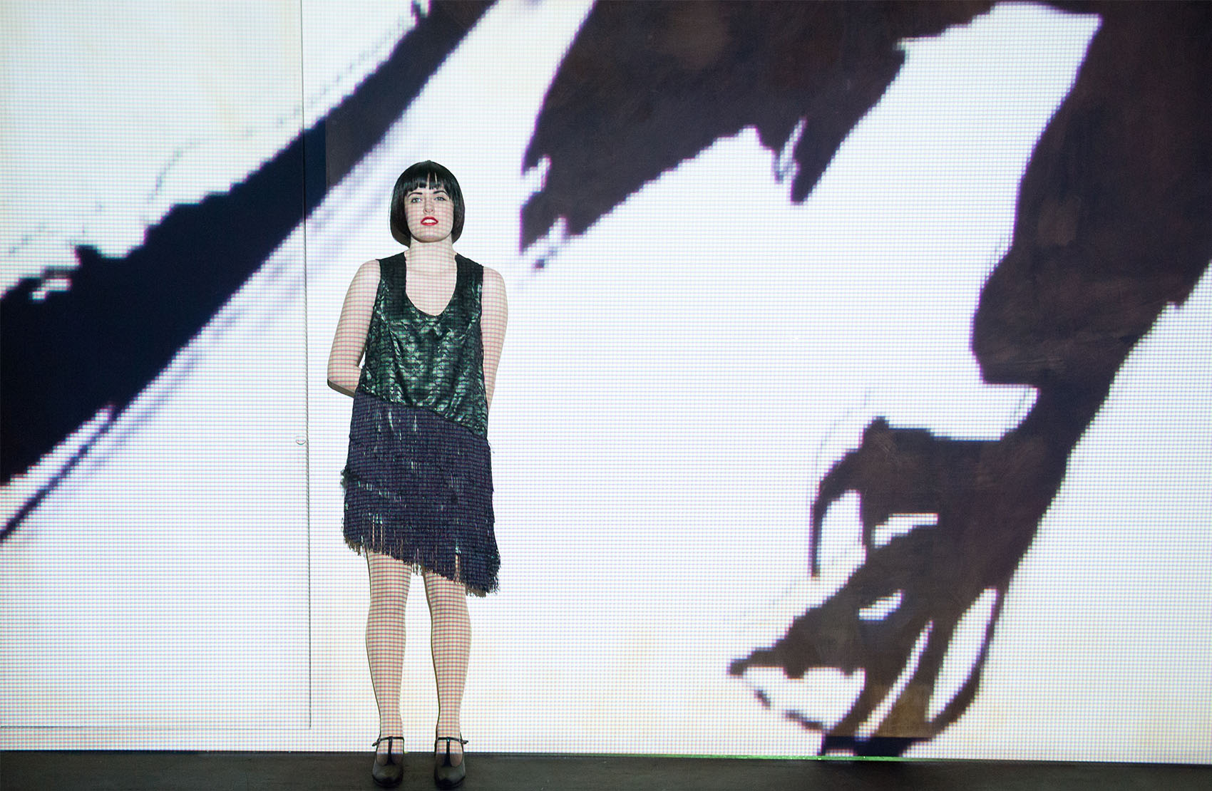 A young white woman in a shiny deep green and black tassel dress dons a black pixie cut stands in front of a brightly lit screen with a large raven projected onto it. 
