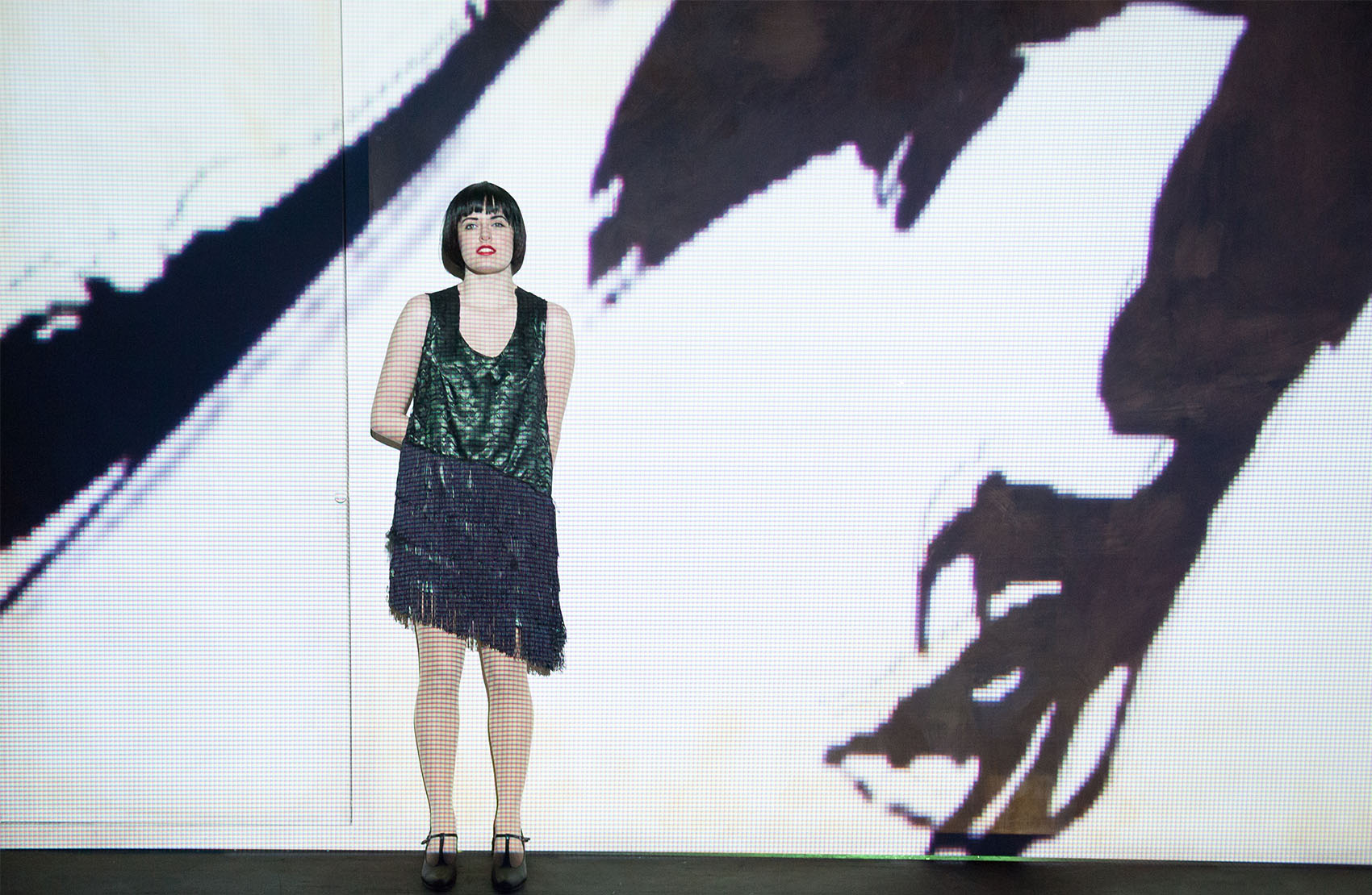 A young white woman in a shiny deep green and black tassel dress dons a black pixie cut stands in front of a brightly lit screen with a large raven projected onto it.