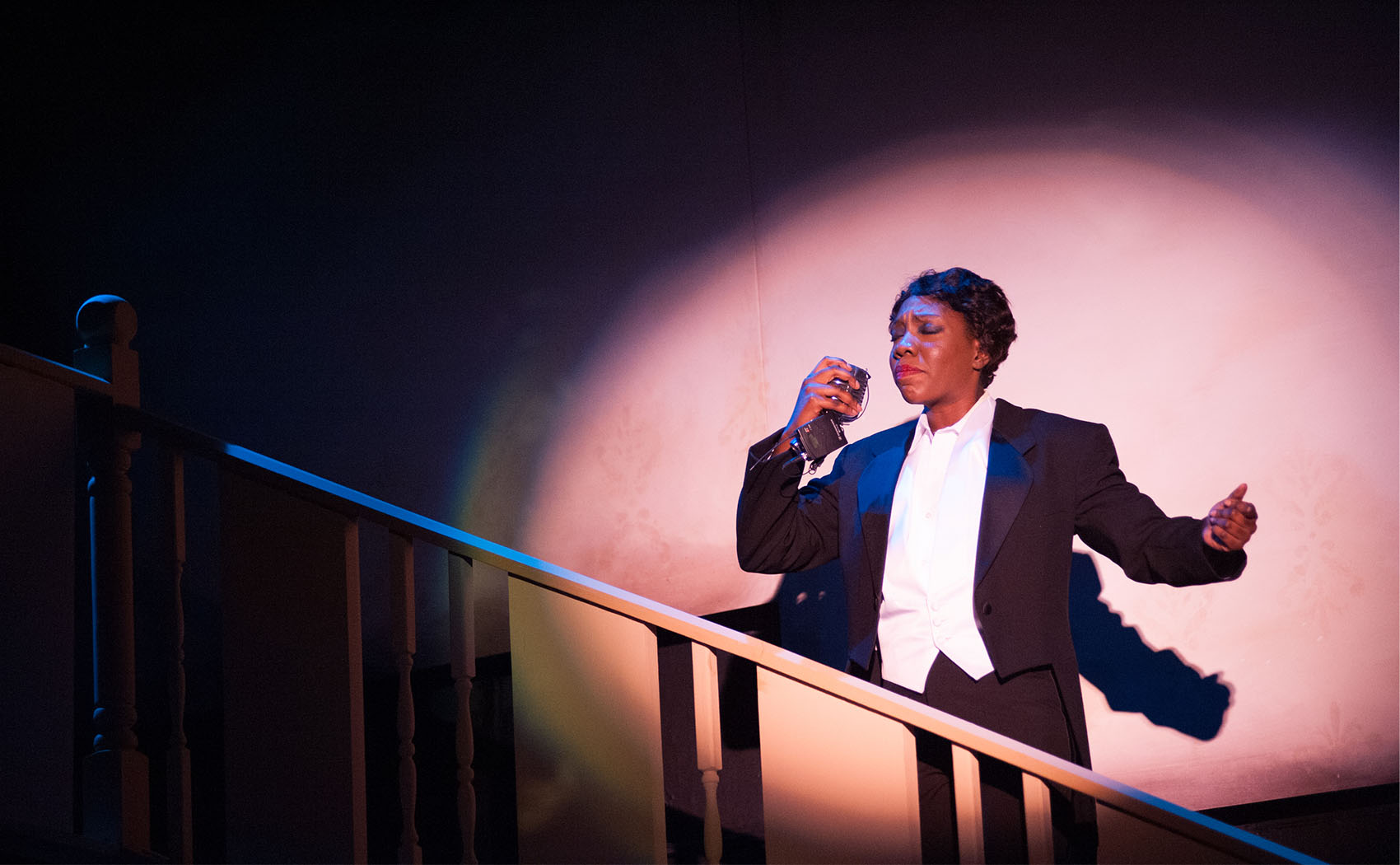 A young black woman sings, eyes closed as she hums a note. She’s dressed in a black and white tuxedo and holds an olden microphone.