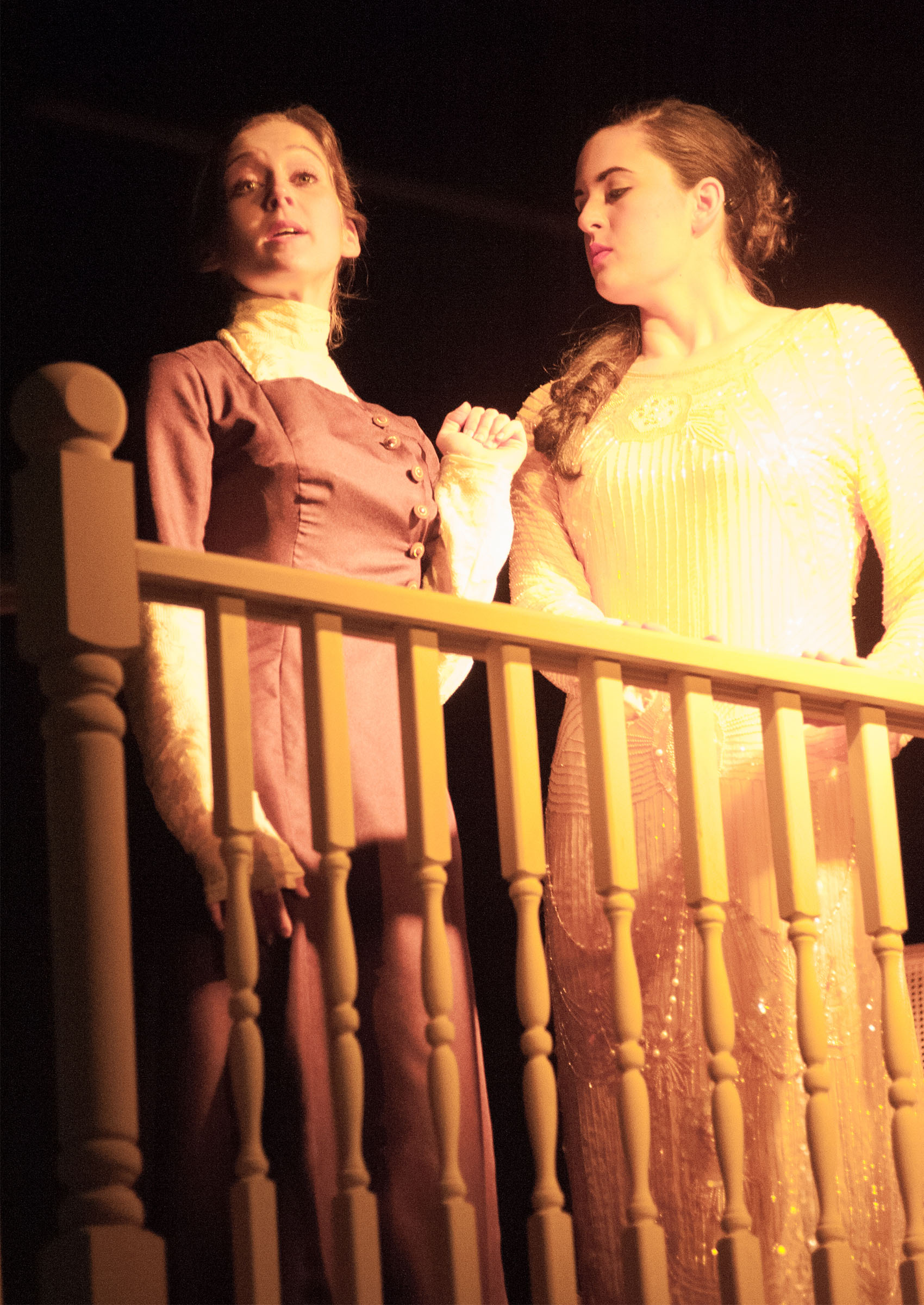 Two women are in conversation, one looking to the audience n=and speaking, the other standing beside her to her left, intently listening and glancing down. They’re standing on a staircase/balcony of sorts, and harsh yellow lighting is cast upon them both.