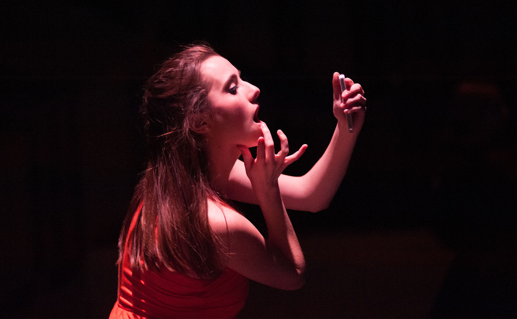 This is a closeup of a young woman staring into a compact mirror, her hand in a motion to fix her lipstick with her middle finger, her thumb pressed under her chin as to hold it up. Lighting is cast on her from above. 