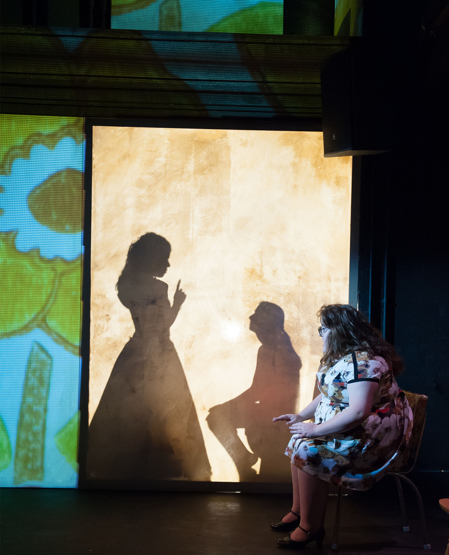 A young woman sitting in a chair looks tensely at a shadow screen, behind it are the silhouettes of a young woman in a dress, a finger to her lip in a hushing motion. The person she is hushing sits and looks up at her, their hair is held back in a ponytail.  Colorful screens are projected beside them. 