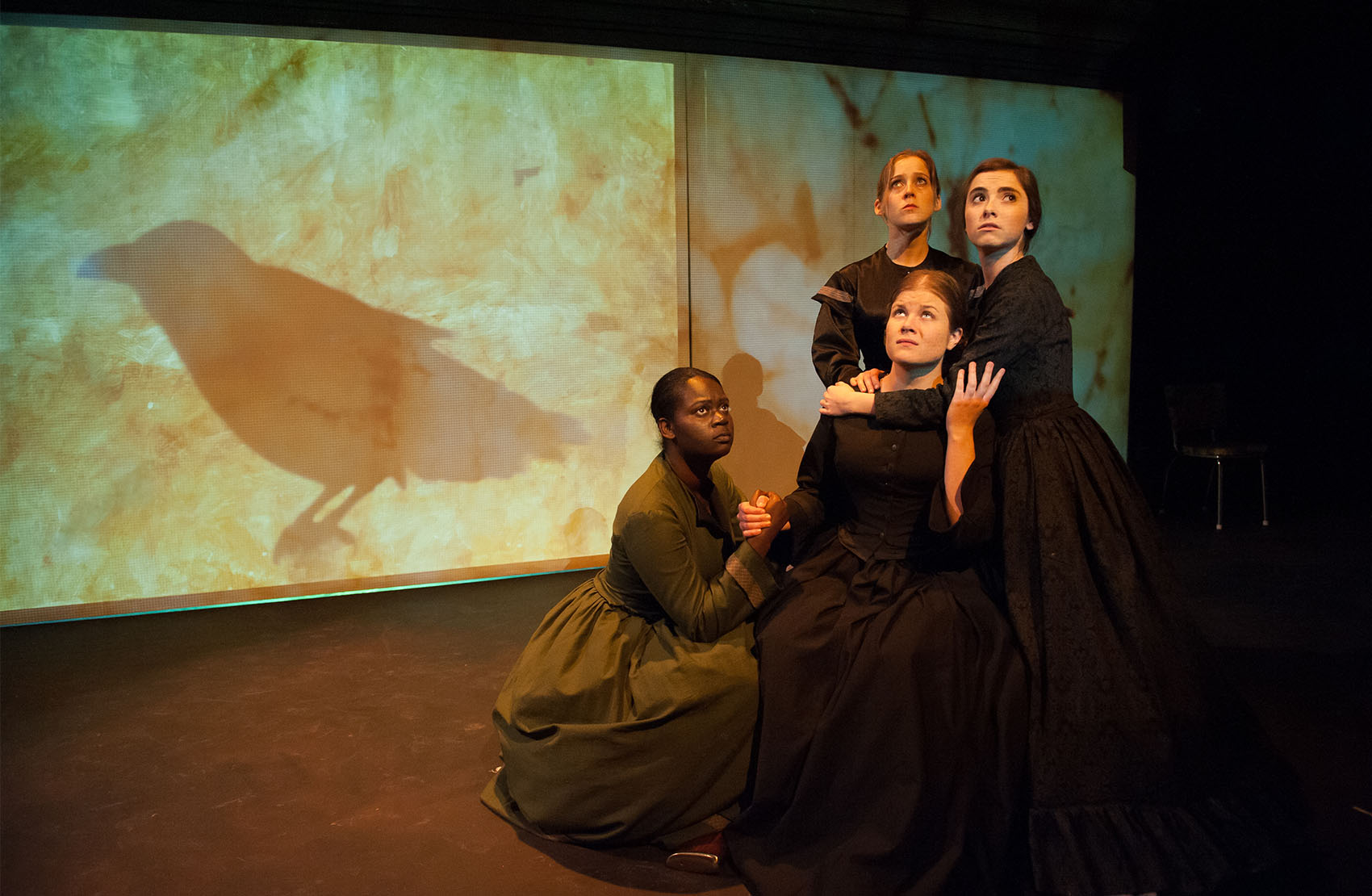 Four young women (one black, 3 white) are huddled near each other. The first woman is in a green vintage dress, crouched next to the 3 other women, who are dressed in all black dresses. They all look up worriedly, a projected raven silhouette on a screen behind them. 