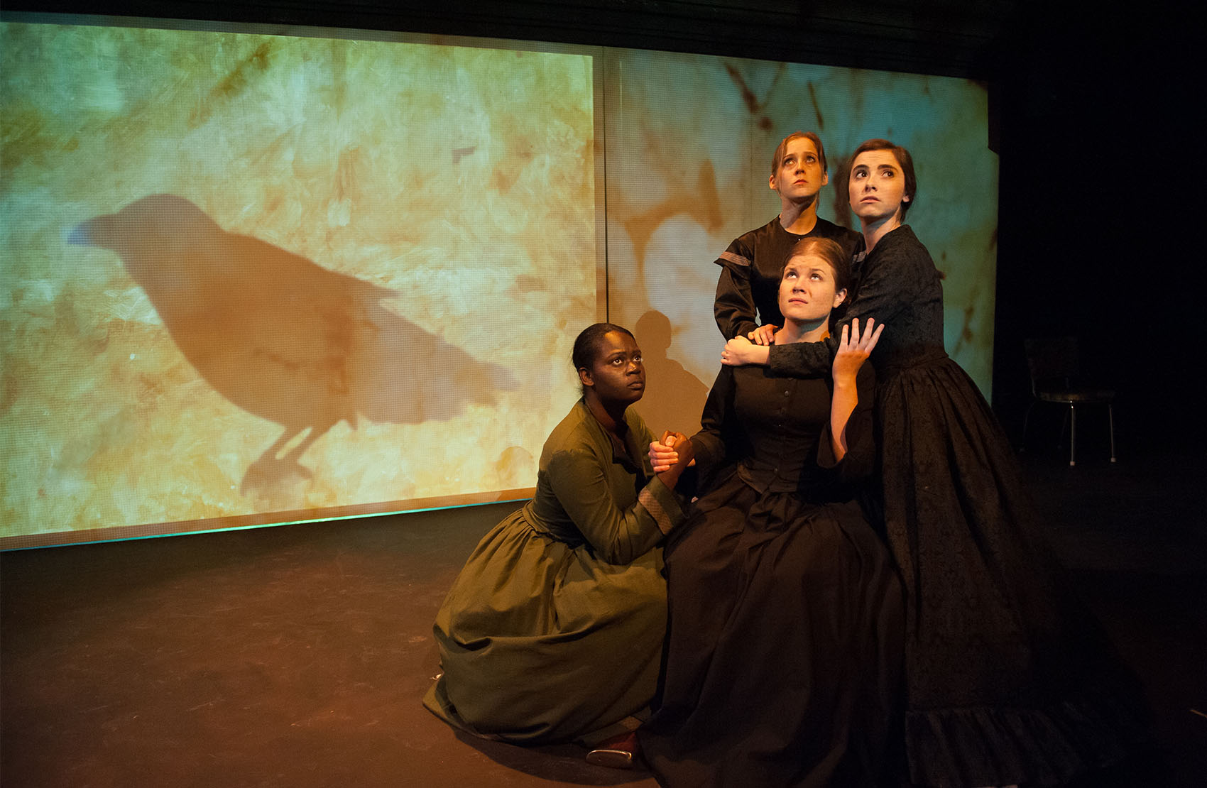 Four young women (one black, 3 white) are huddled near each other. The first woman is in a green vintage dress, crouched next to the 3 other women, who are dressed in all black dresses. They all look up worriedly, a projected raven silhouette on a screen behind them.