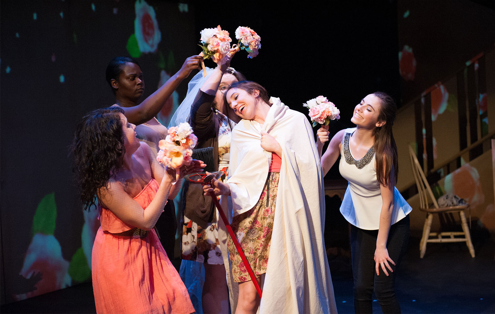 Four young women (1 black, 1 Hispanic, and three white) smile and cheer around one woman, adorning her with flower bouquets, the central woman wrapped in a white cloth as she holds a pair of sunglasses and a red leash.  