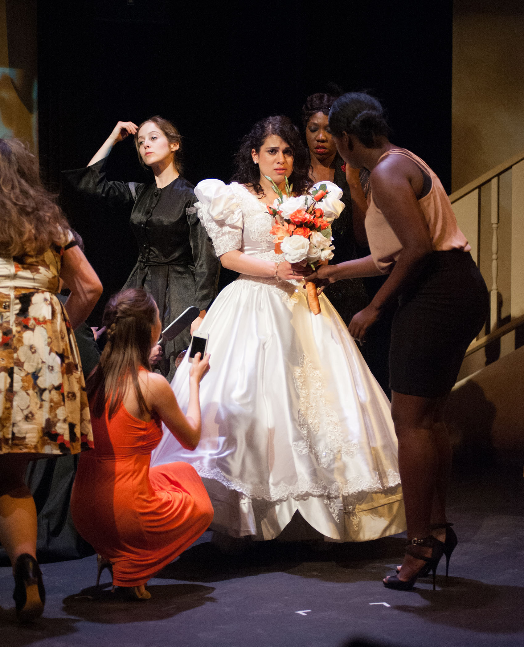 A young Hispanic woman stands centrally in a white wedding dress and holds a bouquet of flowers, looking apprehensive and worried, tears in her eyes. Five other women stand around her, adjusting either themselves or her.  