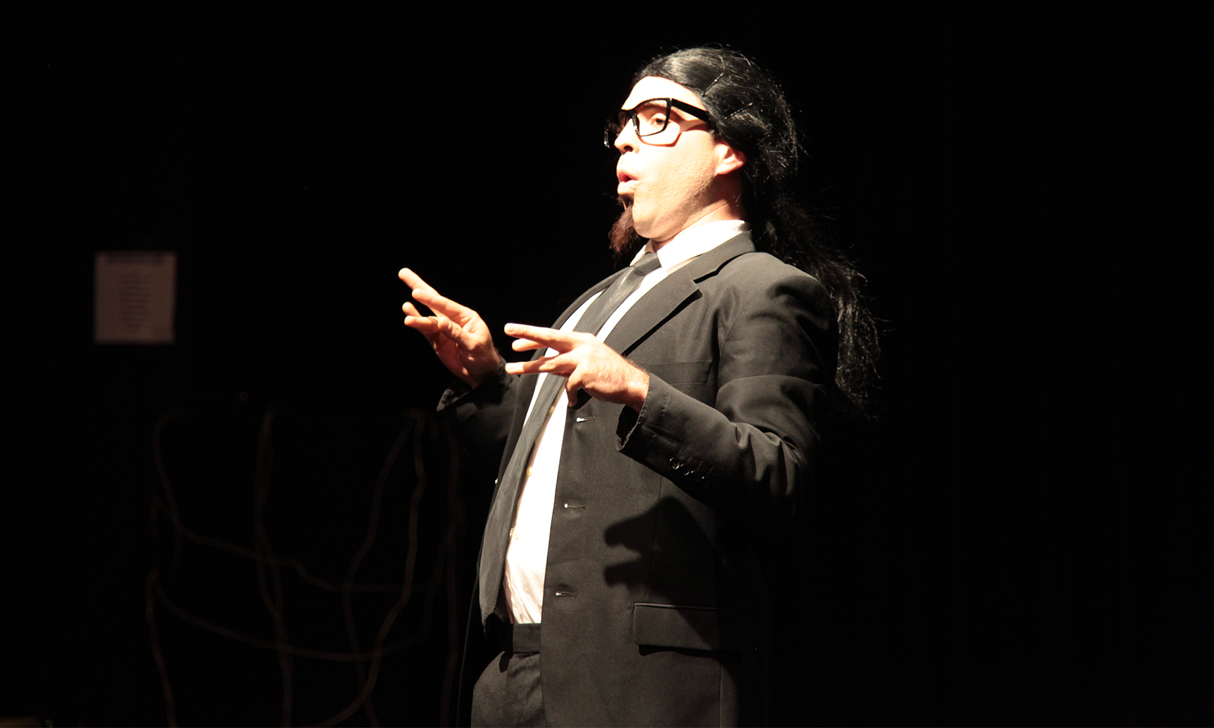 A man with slick black hair and a taped-on goatee is decked in a black and white suit and dons' black glasses has his fingers crookedly outreached as he speaks, he’s making an O shape with his mouth.  