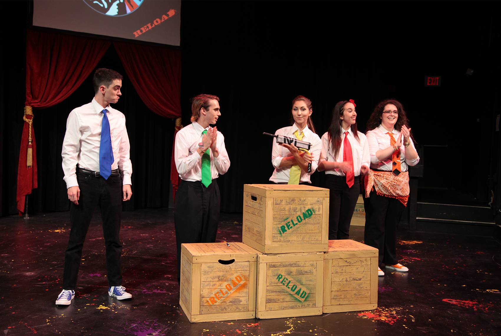 A group of young men and women stand behind some stacked wooden crates that read “RELOAD”. Each person is wearing a white button down with black slacks with a different colored tie ranging from blue (far right), green, yellow, red to orange (far left). A girl with a yellow tie (middle) holds up a makeshift weapon. 