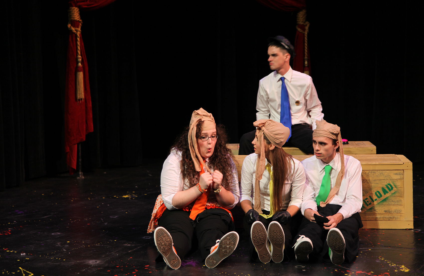 A small group of young men and women are huddled together off to the audience’s left (viewer’s right). Three of them (Orange, Yellow and Green) sit in the foreground in front of crates that read “RELOAD”, pantyhose stockings tied to their heads, each looking noticeably worried. Blue sits behind them on a crate looking to the left.  