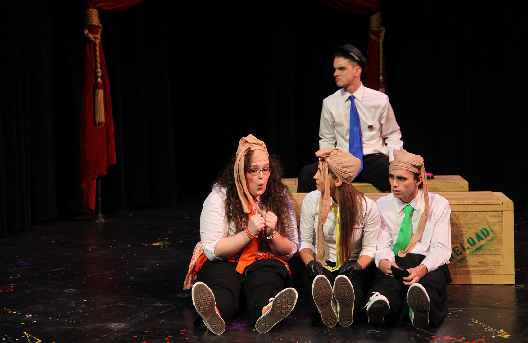 A small group of young men and women are huddled together off to the audience’s left (viewer’s right). Three of them (Orange, Yellow and Green) sit in the foreground in front of crates that read “RELOAD”, pantyhose stockings tied to their heads, each looking noticeably worried. Blue sits behind them on a crate looking to the left.