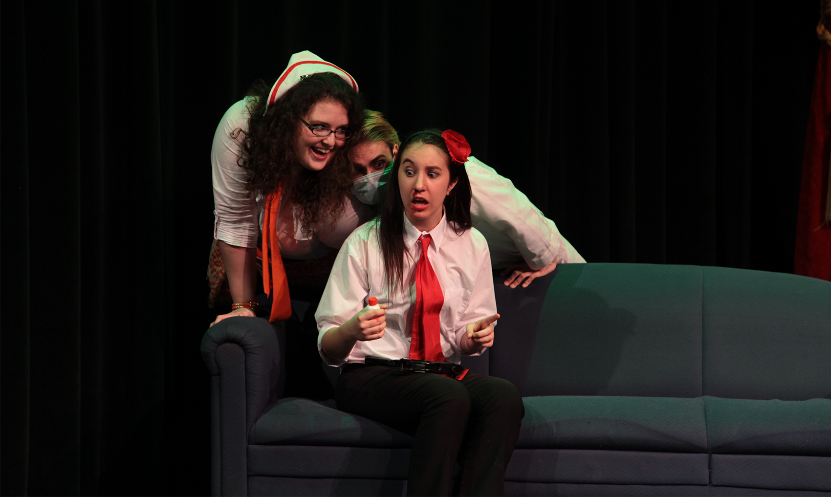 A young woman sits on a couch, with a red tie. Looming behind her is a girl with an orange tie and a young man with a face mask on. They both look intensely at the young woman with the red tie who is startled and looks afraid. Her nose is bleeding and she holds a small white vial with a red cap.