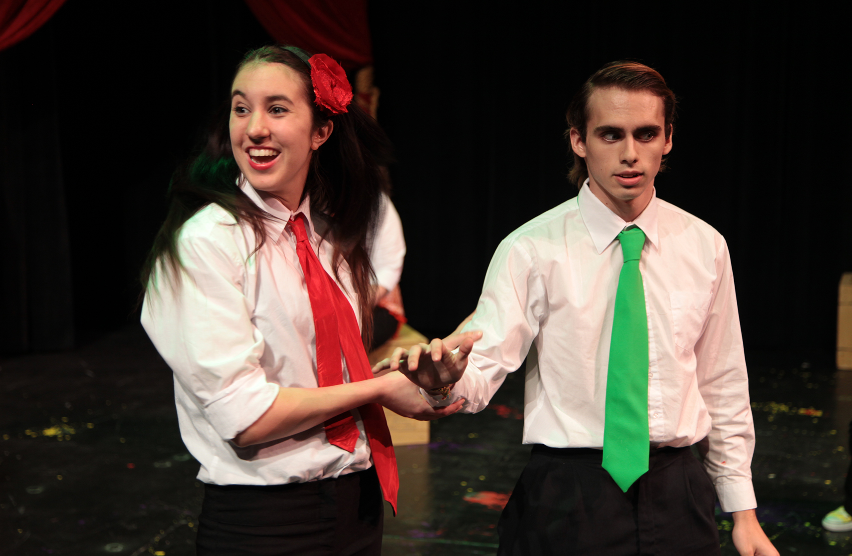 This is a close shot of a young man and woman standing together, the woman in a red tie, holding onto the arm of the man (who wears a green tie). She looks to the left excitedly, the young man looks to the right hurriedly, anxious and seemingly tense.
