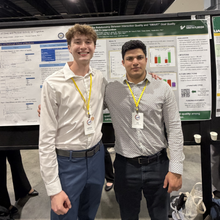 two young men arm around eachother shoulders standing in front of research poster.