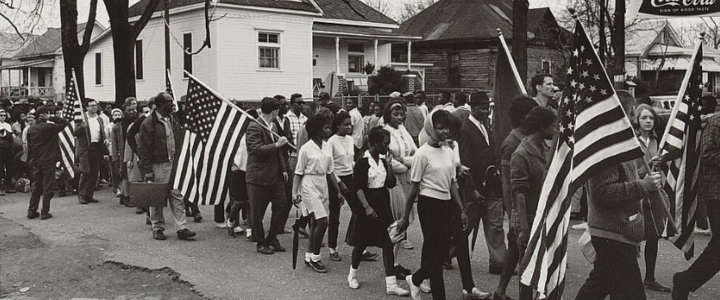 A group of people, several carrying American flags, walk past homes as part of the march from Selma to Montgomery in 1965. The photo is in black and white.