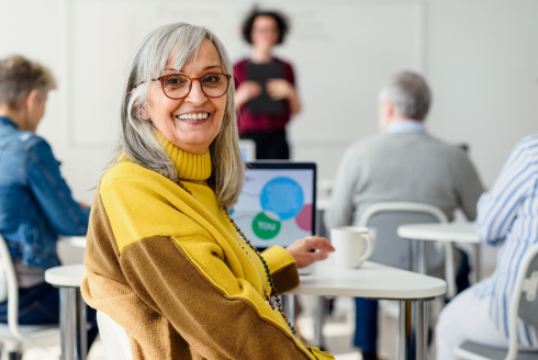 An adult woman wearing a bright yellow sweater and jacket smiles at the camera. A classroom with students and computers is in the background.