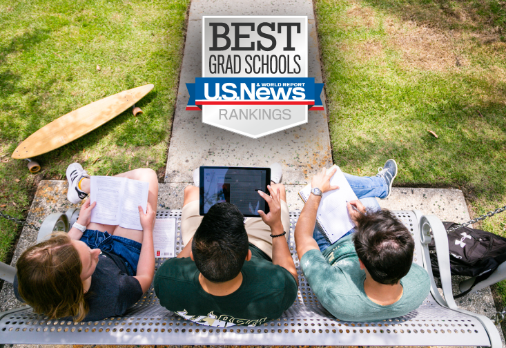 A view from above as three students sit on a bench. One student is reading a book, another is viewing a laptop and the third student is looking at a notebook.