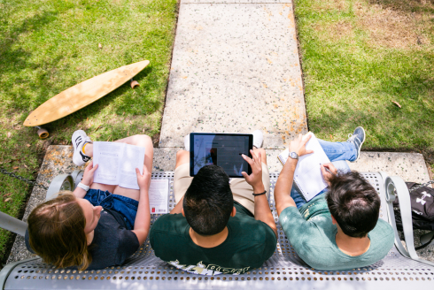 A view from above as three students sit on a bench. One student is reading a book, another is viewing a laptop and the third student is looking at a notebook.
