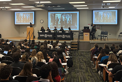 Women in policing panel speak on stage with presentation in the background and students listening to the speakers